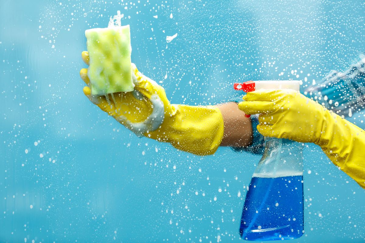 A person wearing yellow rubber gloves holds a spray bottle, ready to clean or disinfect a surface.
