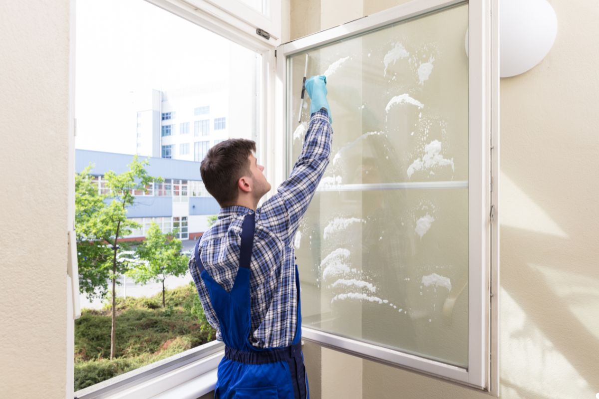 A man in blue overalls is cleaning a large window with a squeegee, focused on his task.