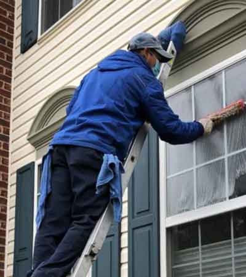 A man cleaning house windows from a ladder, showcasing effective window cleaning practices for homeowners.